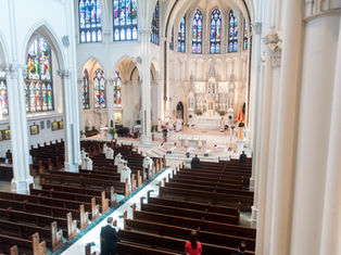 Wide view of a cathedral interior with stained glass windows. Clergy in robes perform a ceremony at the altar, while people observe from pews.