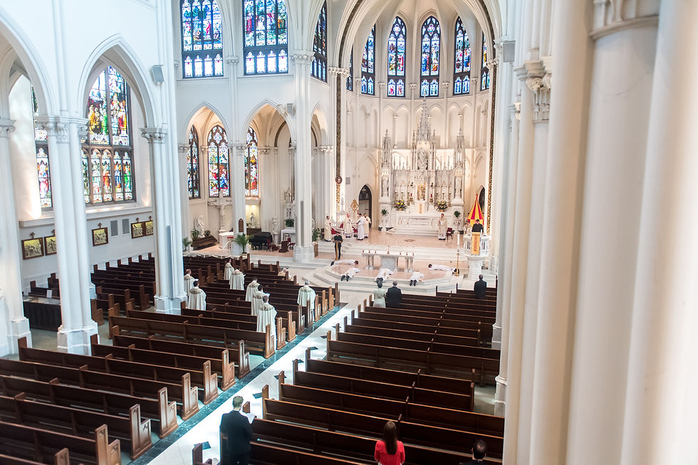 Wide view of a cathedral interior with stained glass windows. Clergy in robes perform a ceremony at the altar, while people observe from pews.