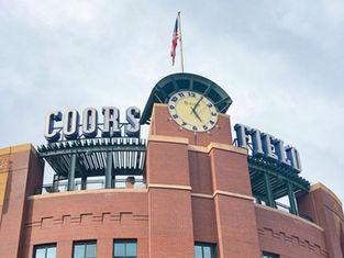 The sign above Coors Field baseball field in Denver stands out against a cloudy sky. An American flag waves in the wind above the sign.
