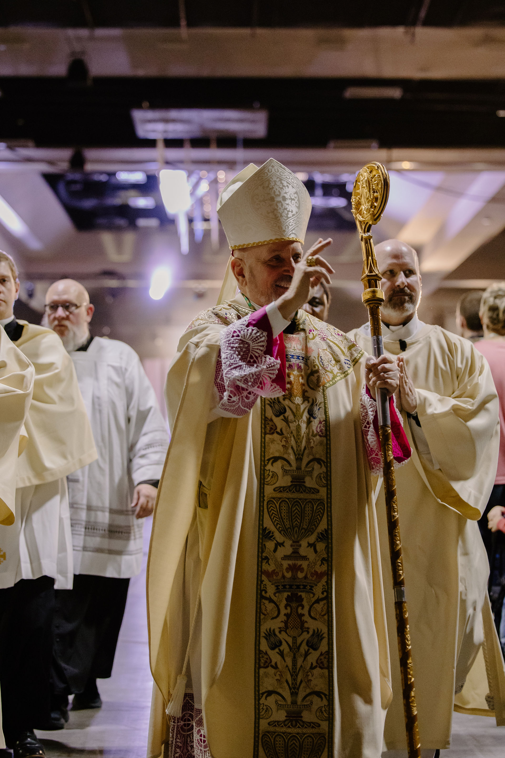 A bishop in ornate robes and mitre gives a blessing, holding a gold staff. Clergy in cream attire stand nearby, inside a grand hall.