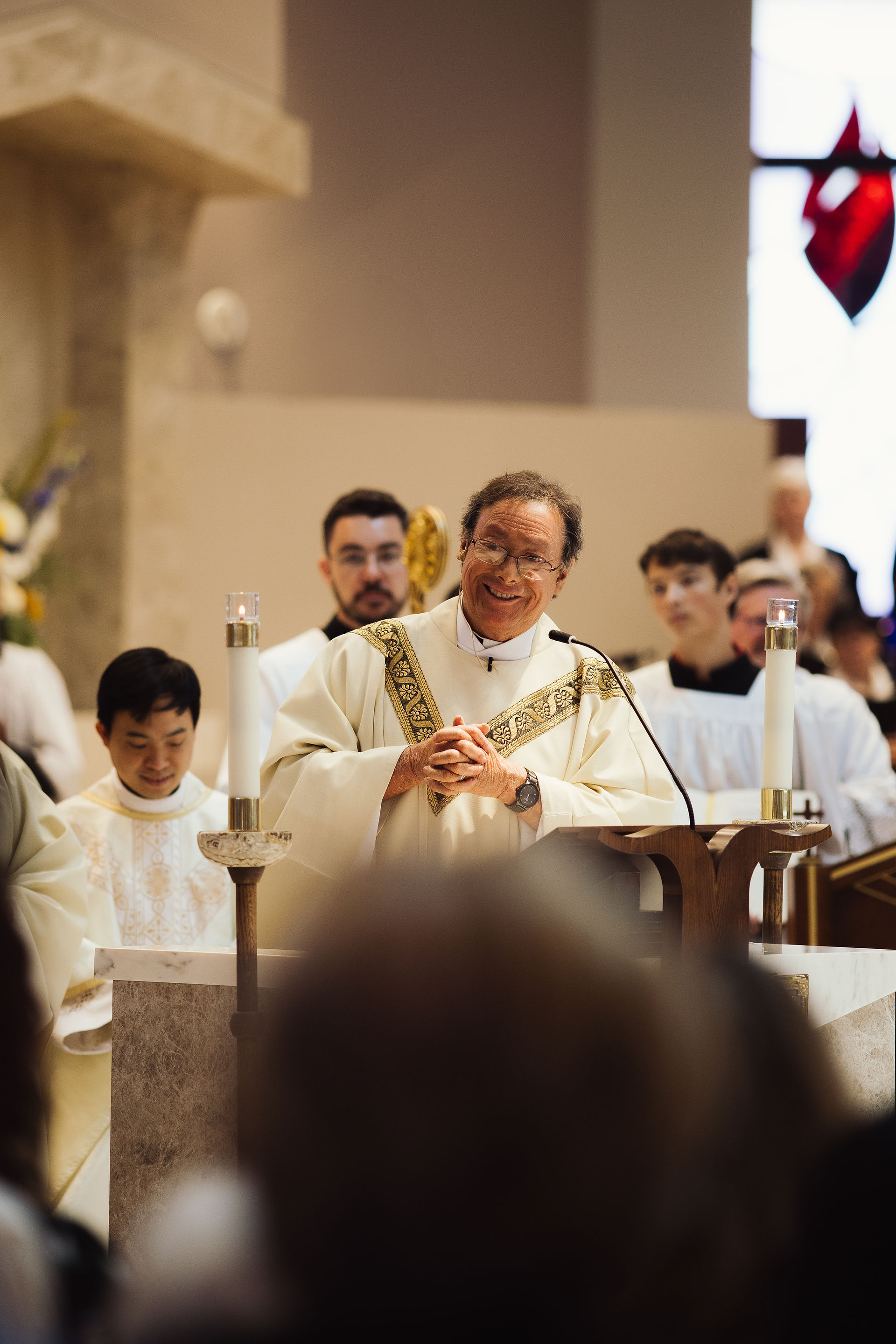 Priest smiling at a pulpit, surrounded by congregation in a church. Bright interior, man in white robe, candles on altar. Emotive atmosphere.