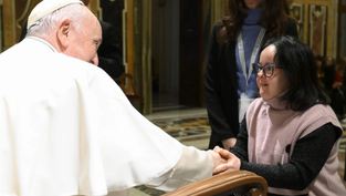 An elderly man in white robes shakes hands with a woman in a pink sweater in an ornate room, conveying warmth and connection.