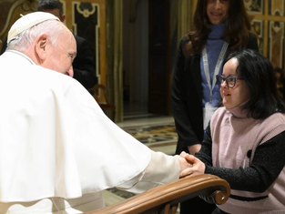 An elderly man in white robes shakes hands with a woman in a pink sweater in an ornate room, conveying warmth and connection.