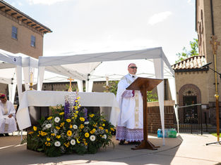 Bishop in white vestments speaks at outdoor altar under white canopy, with flowers and church buildings behind him.