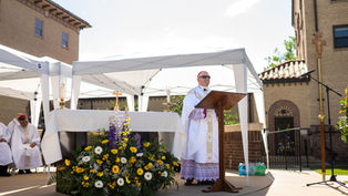 Bishop in white vestments speaks at outdoor altar under white canopy, with flowers and church buildings behind him.