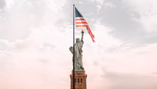 The Statue of Liberty with the American flag flies against a cloudy sky. Soft pink and gray hues create a serene and patriotic atmosphere.