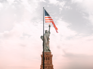 The Statue of Liberty with the American flag flies against a cloudy sky. Soft pink and gray hues create a serene and patriotic atmosphere.