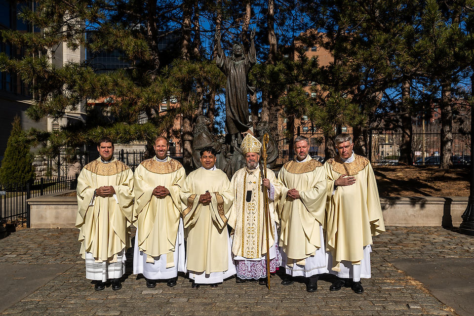 Six clergy in ornate cream robes stand on a cobblestone path, with a statue behind them and trees overhead, under clear blue skies.