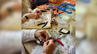 Hands make beaded jewelry at a craft table with pliers, beads, trays, and colorful tools on towels.
