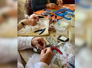 Hands make beaded jewelry at a craft table with pliers, beads, trays, and colorful tools on towels.