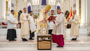 Clergy in white and gold robes conduct a religious ceremony in a grand cathedral. One holds a book, another pours oil. The mood is solemn.