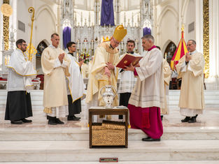 Clergy in white and gold robes conduct a religious ceremony in a grand cathedral. One holds a book, another pours oil. The mood is solemn.