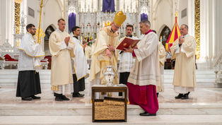 Clergy in white and gold robes conduct a religious ceremony in a grand cathedral. One holds a book, another pours oil. The mood is solemn.