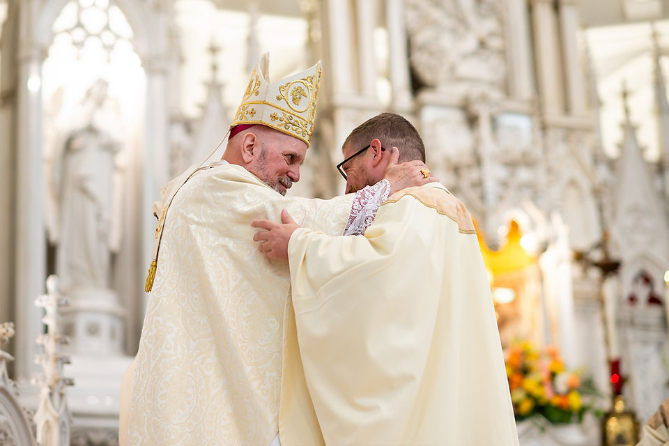Two clergymen in ornate robes embrace warmly in a detailed church setting. Ornate white decor and colorful flowers in the background.