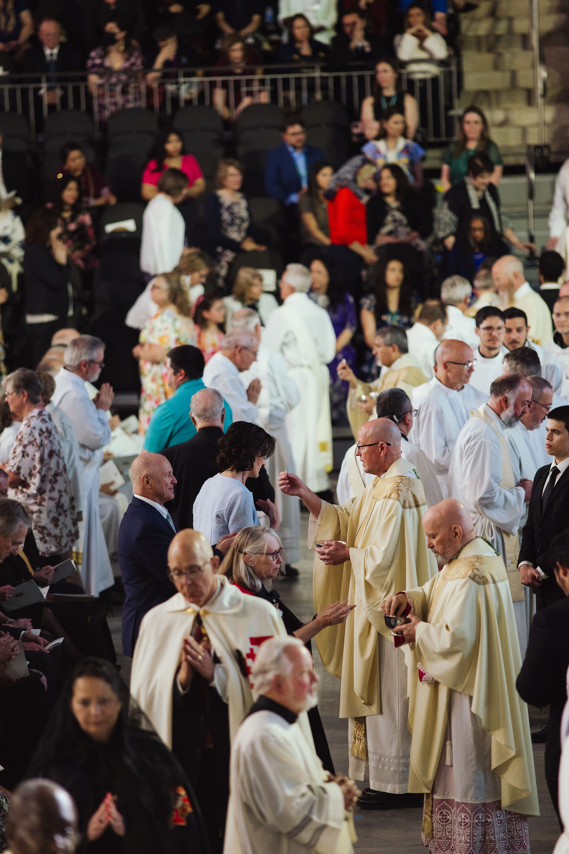 Priests distribute Communion to a large congregation gathered in a crowded indoor arena during a Catholic Mass.