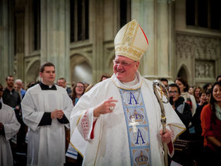 A bishop in ornate robes and miter smiles, holding a staff. Clergy in white robes follow. Cathedral setting, people in background smiling.