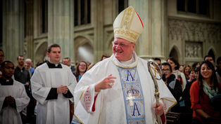 A bishop in ornate robes and miter smiles, holding a staff. Clergy in white robes follow. Cathedral setting, people in background smiling.