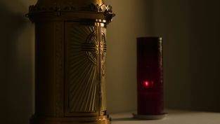Golden tabernacle with engraved cross beside a glowing red candle in a dimly lit setting, creating a serene and reverent atmosphere.