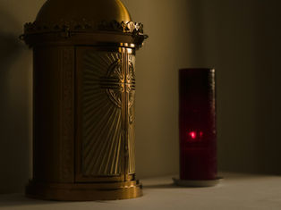 Golden tabernacle with engraved cross beside a glowing red candle in a dimly lit setting, creating a serene and reverent atmosphere.