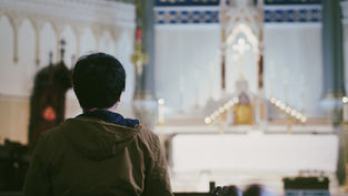 Person in a brown jacket sitting in a church pew, facing an ornate altar with candles and cross, under a serene atmosphere.