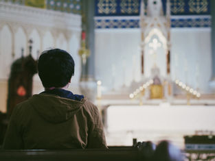 Person in a brown jacket sitting in a church pew, facing an ornate altar with candles and cross, under a serene atmosphere.