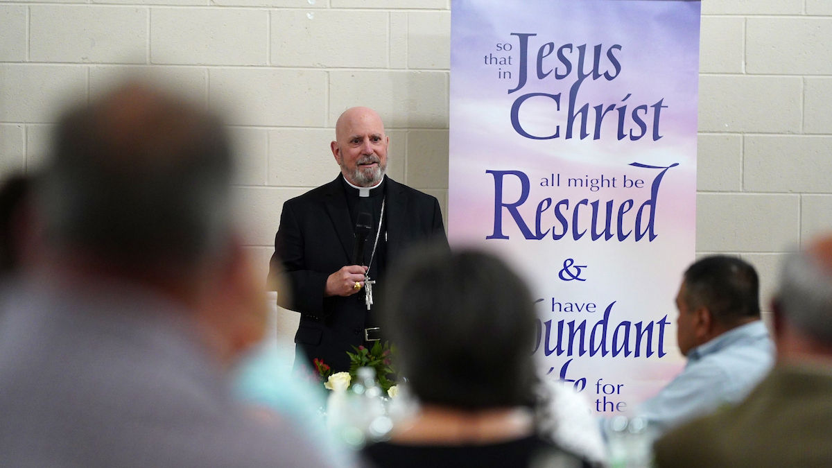 Bald man in black clergy attire speaks at an event, standing by a banner with Christian text. Attendees sit in foreground. Neutral mood.