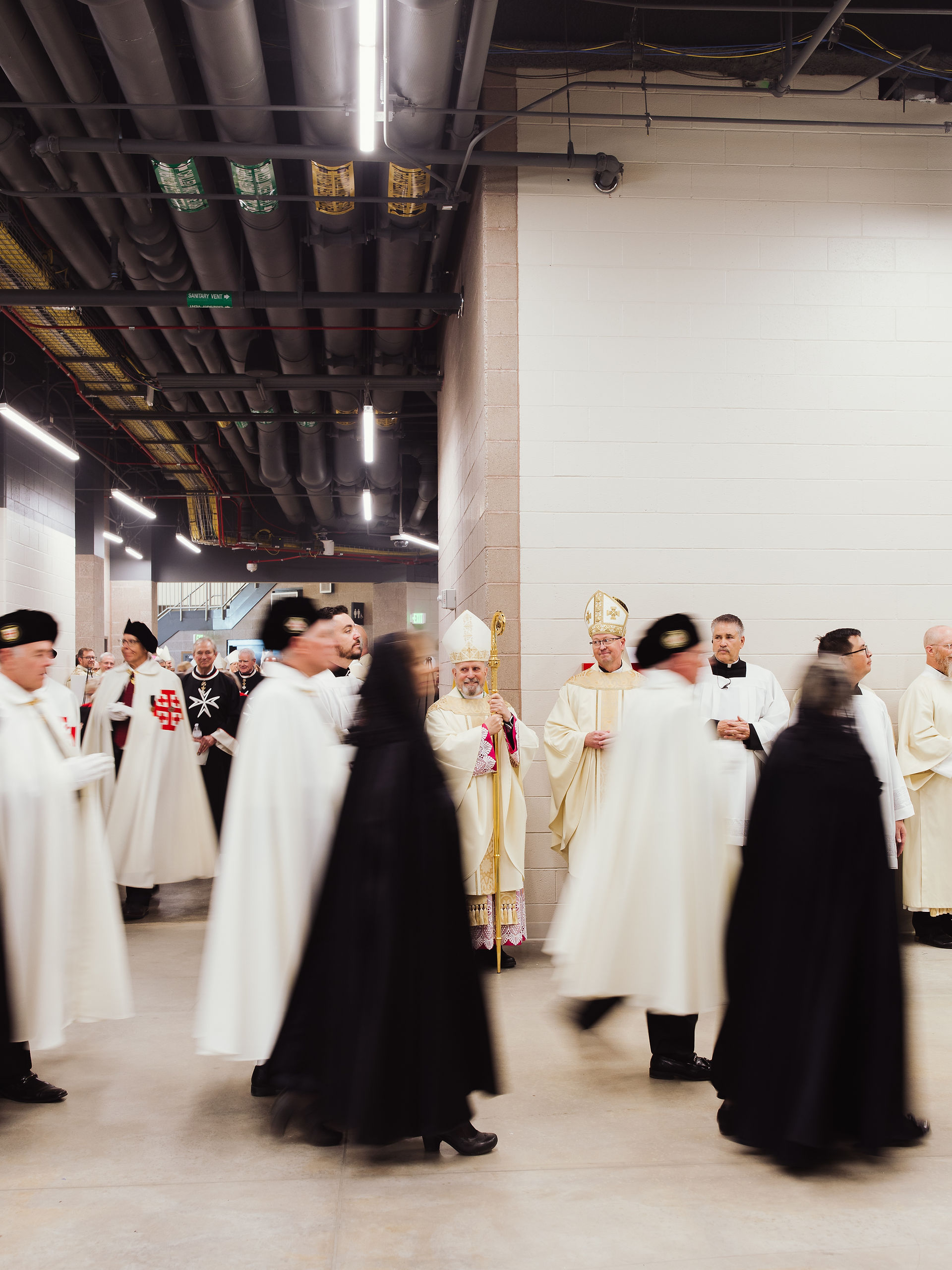 People in white and black robes walk in a hallway. Clergy in the background, one holds a staff. Ceiling pipes visible. solemn atmosphere.
