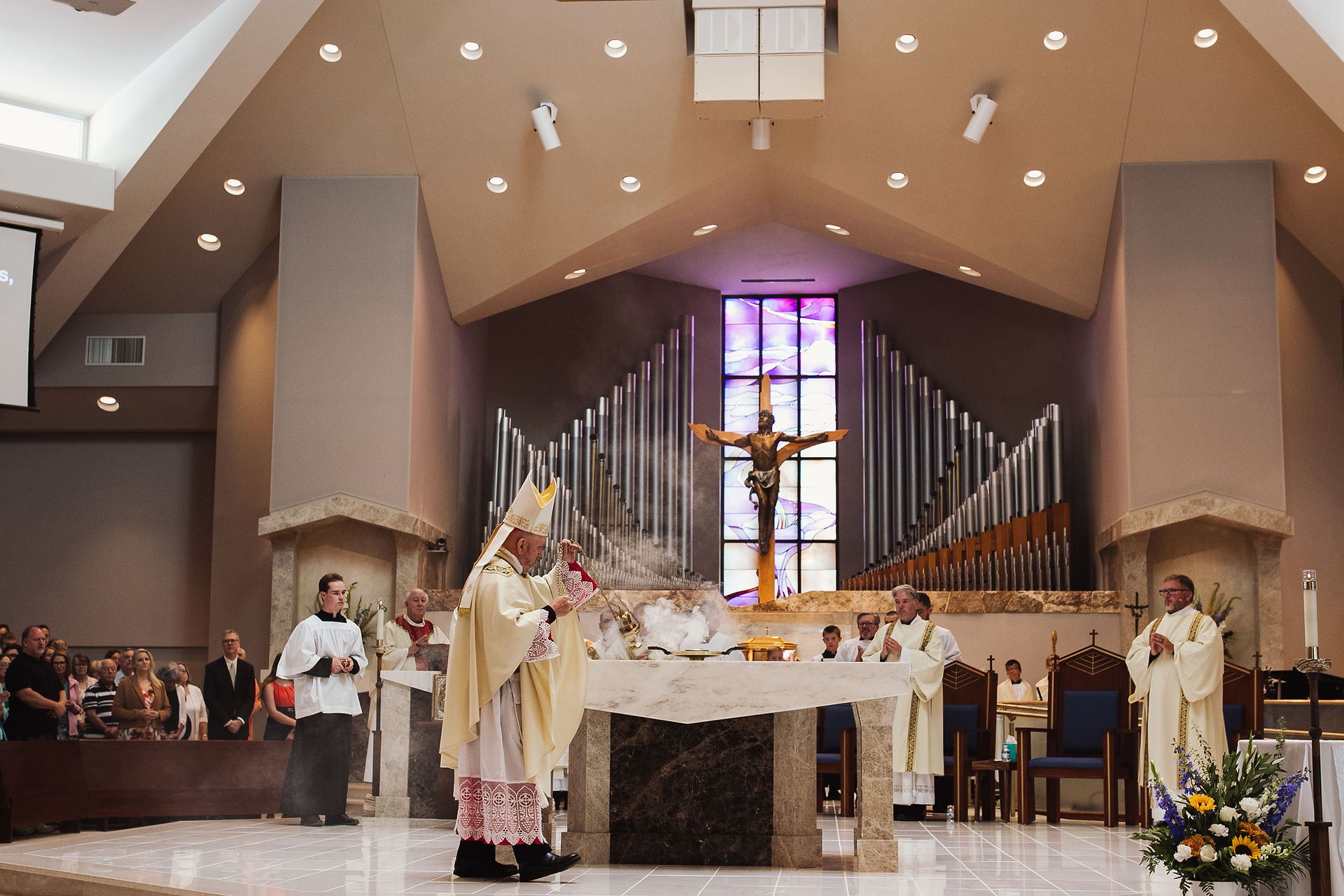 A bishop in ornate robes swings a censer, releasing smoke in a church. Behind, a crucifix and organ pipes; congregants watch solemnly.