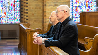 Two Catholic bishops sit in wooden pews inside a church, hands folded in prayer, with stained glass windows behind them.