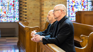 Two Catholic bishops sit in wooden pews inside a church, hands folded in prayer, with stained glass windows behind them.