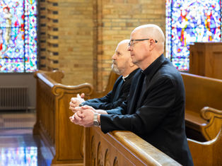 Two Catholic bishops sit in wooden pews inside a church, hands folded in prayer, with stained glass windows behind them.