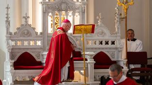Bishop in red vestments prays at an ornate white altar in a church, with altar servers nearby and warm golden light.