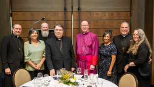 A group of eight people, including clergy and others, smile at a dinner event. They're in formal attire against a backdrop with symbols.