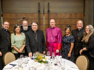 A group of eight people, including clergy and others, smile at a dinner event. They're in formal attire against a backdrop with symbols.