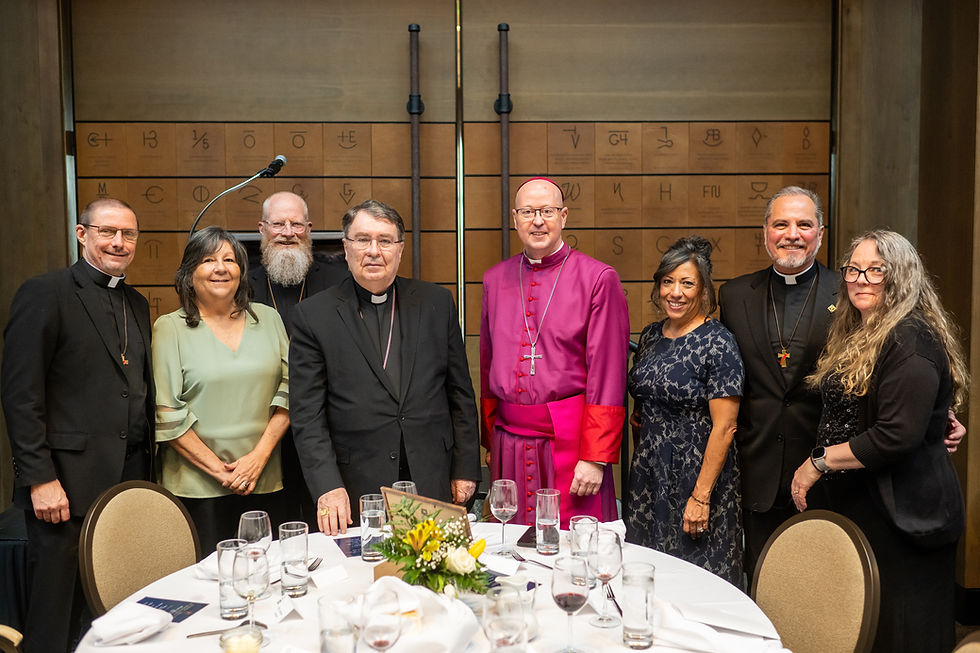 A group of eight people, including clergy and others, smile at a dinner event. They're in formal attire against a backdrop with symbols.