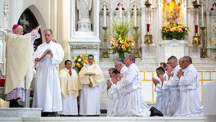 Men in white robes kneel in prayer, while a bishop in ornate robes blesses them in a richly decorated church with floral arrangements.