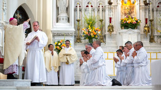 Men in white robes kneel in prayer, while a bishop in ornate robes blesses them in a richly decorated church with floral arrangements.