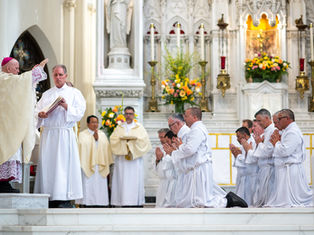 Men in white robes kneel in prayer, while a bishop in ornate robes blesses them in a richly decorated church with floral arrangements.