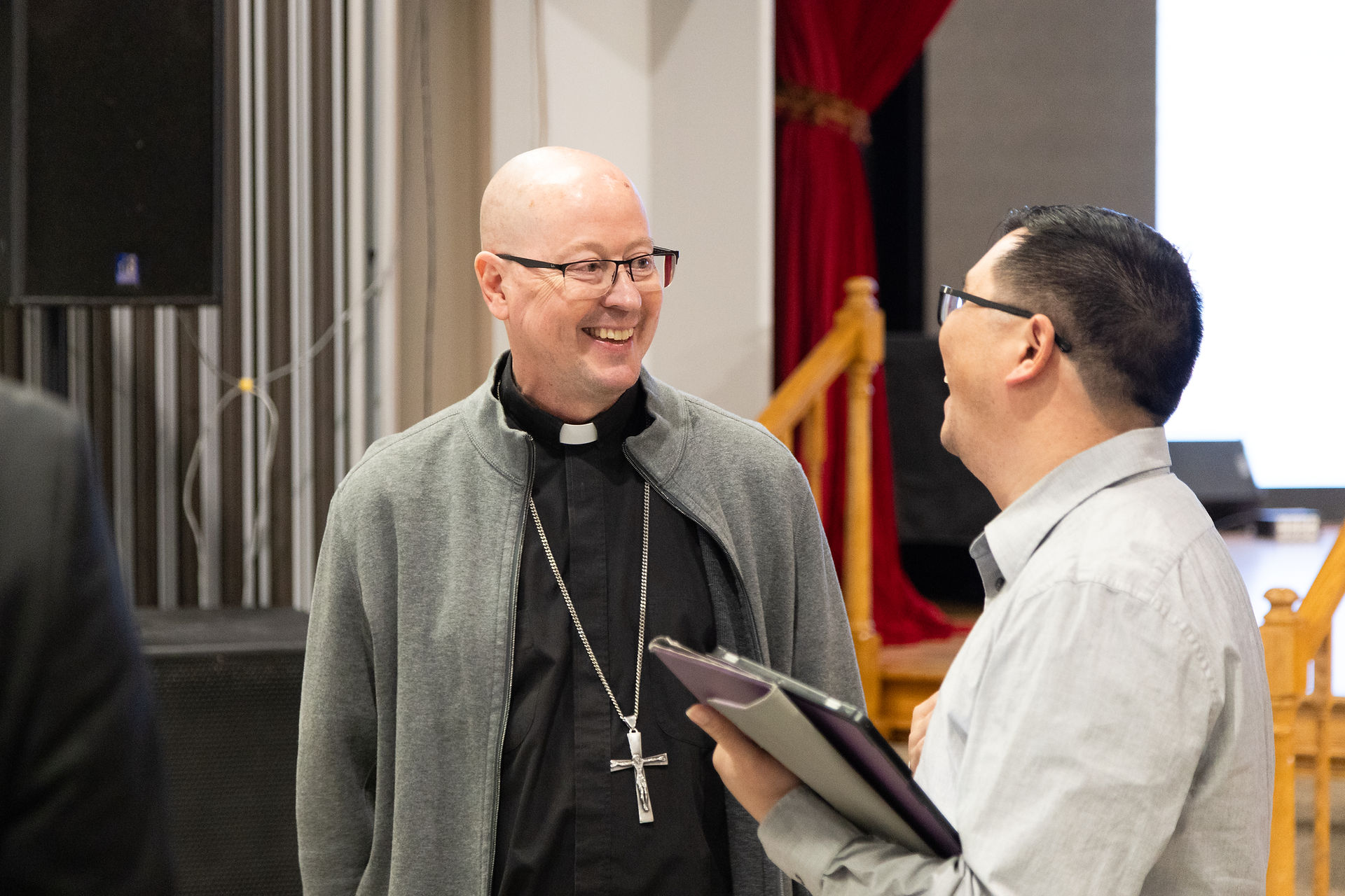 Two men smiling and talking indoors, one in a clerical collar, the other holding a folder. Background shows a red curtain and wooden steps.