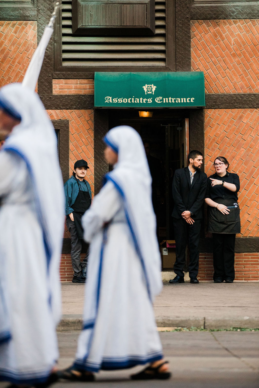 Blurred nuns walk past a brick building with an Associates Entrance sign, while two staff stand talking at the doorway.