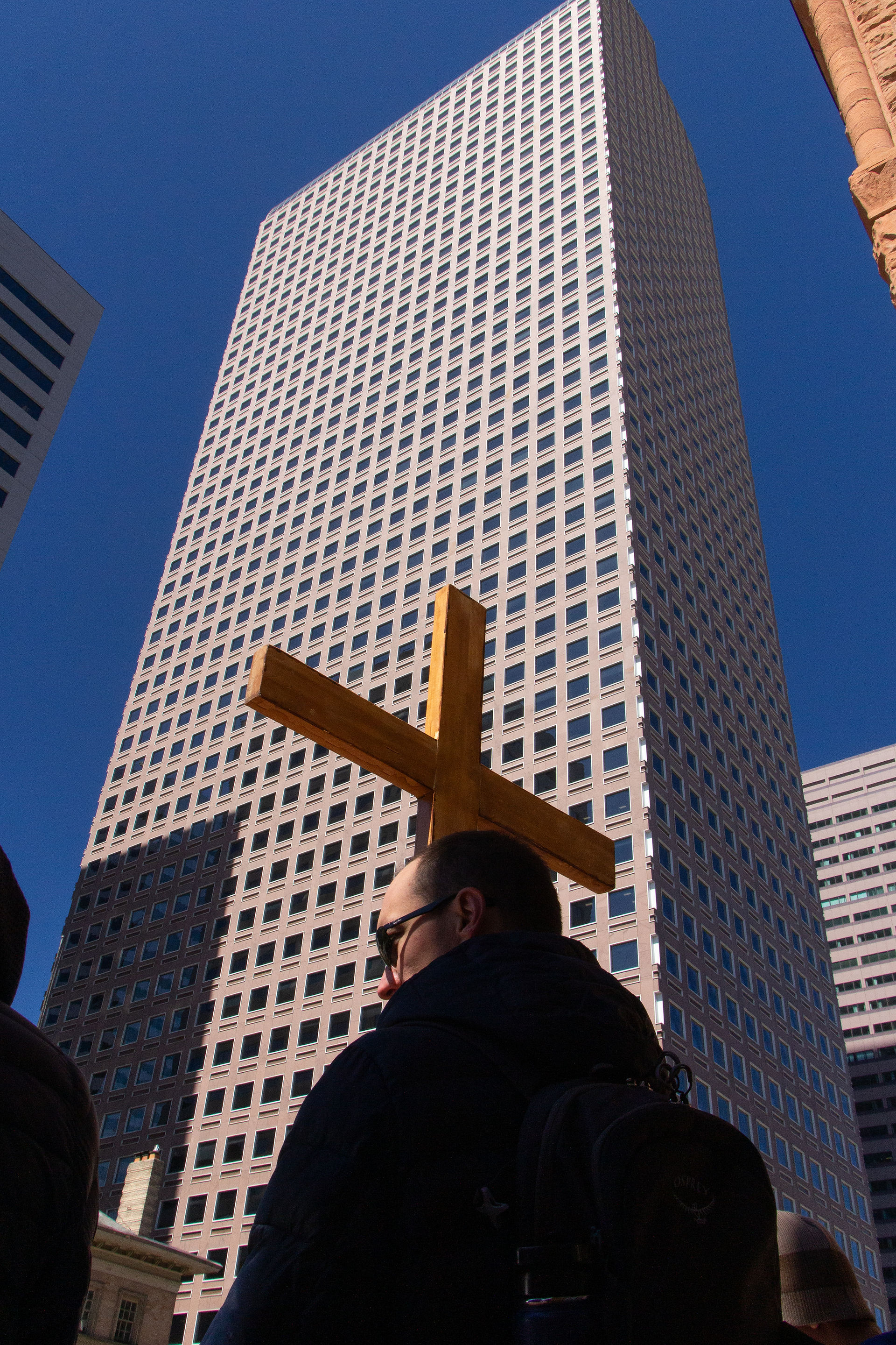 A wooden cross is held in front of a tall city skyscraper during an outdoor Way of the Cross procession.