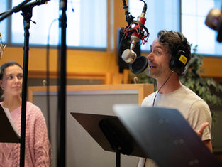 Two people in a recording studio, speaking into microphones. One wears a pink sweater, the other a white shirt and headphones. Bright room.
