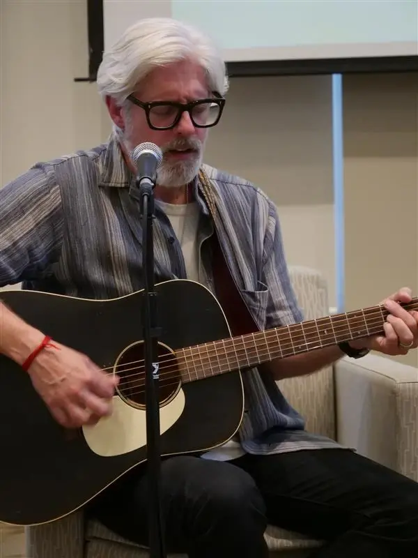 A man with white hair in a blue shirt playing a black guitar.