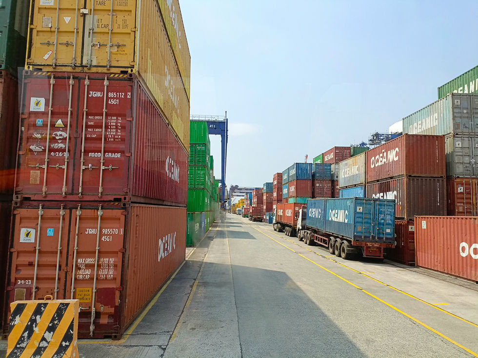 Stacked shipping containers in various colors line a port with trucks nearby. A clear sky background and road markings indicate a busy dock.