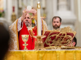 A man in ornate red and gold vestments holds a wafer aloft during a religious ceremony. Elegant altar decor and candle in the foreground.