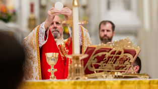 A man in ornate red and gold vestments holds a wafer aloft during a religious ceremony. Elegant altar decor and candle in the foreground.