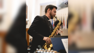 Man in a black suit plays a gold saxophone in a formal hall, with blurred flags behind him.