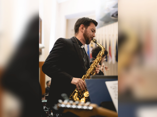 Man in a black suit plays a gold saxophone in a formal hall, with blurred flags behind him.