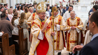 A bishop in ornate robes and mitre holds a staff, leading a procession in a crowded church. Attendees watch attentively.