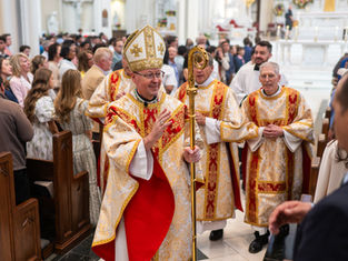 A bishop in ornate robes and mitre holds a staff, leading a procession in a crowded church. Attendees watch attentively.
